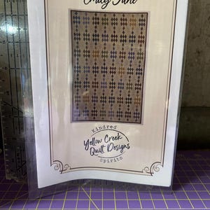 Puede incluir: Un paquete que presenta un patrón de edredón titulado "Diamonds For Emily Jane". El patrón muestra un diseño de diamantes en tonos marrones, azules y morados. El paquete también incluye el logotipo "Yellow Creek Quilt Designs".