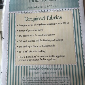 May include: A package for a Patchwork Sunflower Table Runner. The finished size is 31.75 cm x 134.62 cm. The package lists required fabrics, including yellow scraps, green scraps, brown plaid, teal, aqua fabric, and a backing piece.