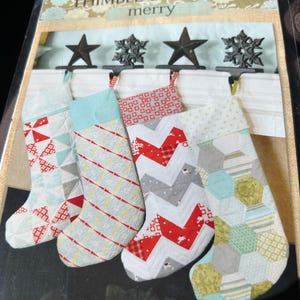 May include: A package of Christmas stocking patterns. The stockings are in various colors and patterns, including red, white, gray, and blue. The package includes the text "THIMBLE BLOSSOMS merry" and the number "#141".