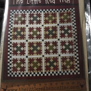 May include: A quilt pattern titled "The Little Red Hen" with a patchwork design. It features red, green, and cream-colored star and cross motifs. The pattern includes the website address www.thelittleredhenonline.com and the name "Ruby Rose".