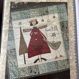 May include: A fabric wall hanging with a folk art design. The central image features a figure in a red dress with the word "JOY" and the text "Peace to All". A Christmas tree and a bird are also depicted. The words "Hatched and Patched" are at the top.