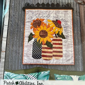 May include: A quilted wall hanging with sunflowers in mason jars. The artwork features the words "Hello Sunshine". The dimensions are 22" x 25". The jars have star, stripe, and solid designs. A ladybug is on one of the sunflowers.