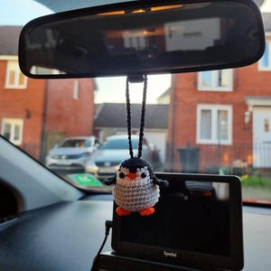 May include: A crocheted penguin car accessory. The penguin has a grey body, black head and wings, orange feet, and a white belly. It is suspended from a car's rearview mirror by a black string.