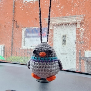 May include: A crocheted grey pigeon car charm with orange feet and beak, and a teal and purple chest. The bird hangs from a dark grey cord, adding a whimsical touch to the car's interior. The background shows a rainy day.