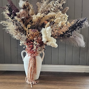 May include: A large, dried floral arrangement in a white vase with handles. The bouquet features a variety of textures and colors, including beige, brown, burgundy, and cream. The vase is placed on a wooden floor against a gray wall.