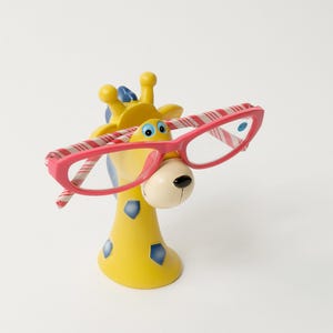 May include: A whimsical yellow giraffe figurine with blue accents and a white face, wearing pink-framed eyeglasses with red and white striped temples. The giraffe has a round, beige nose and is set against a white background.