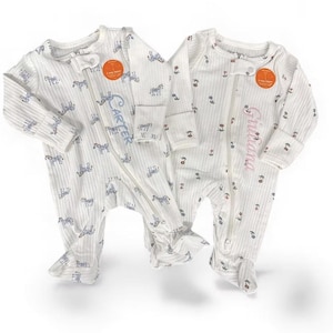 May include: Two white baby footed pajamas with a zebra print and a zipper closure. One pajama has the name "Carter" embroidered in blue, and the other has "Giuliana" embroidered in pink. Both have a round orange tag.
