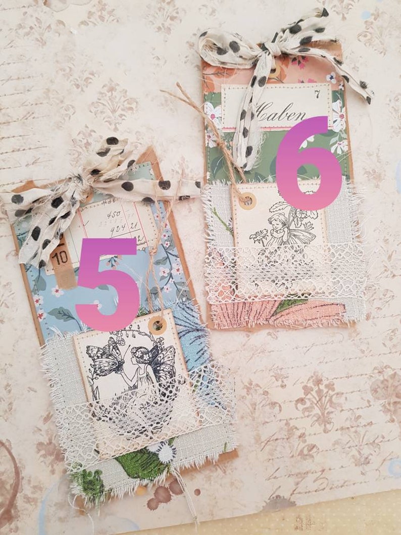May include: Two handmade paper tags with a vintage aesthetic. The tags are decorated with lace, fabric scraps, and floral patterns. The top tag has the number "6" written on it in pink and the word "Calen" written on a smaller tag. The bottom tag has the number "5" written on it in pink. Both tags feature a black and white illustration of a fairy.