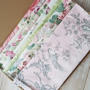 Op de afbeelding: Een set van 10 stofstalen met bloemmotieven in roze, groene en witte tinten. De stoffen zijn gerangschikt in een doos, waarbij de bovenste stof een delicaat bloemmotief met een vogelontwerp heeft.