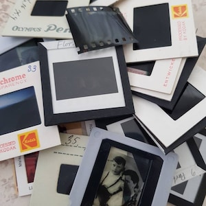May include: A pile of vintage slide film negatives and transparencies, some with black frames and others with white frames. The slides are labelled with various brands, including Olympus, Kodak, and Agfa. Some slides have handwritten notes on them, such as "Flor" and "83".