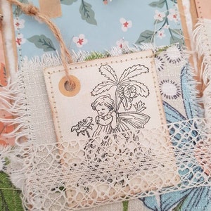 May include: A vintage-style fabric tag featuring a black and white illustration of a girl holding a flower. The tag is decorated with lace and has a brown circle with a hole in the centre.