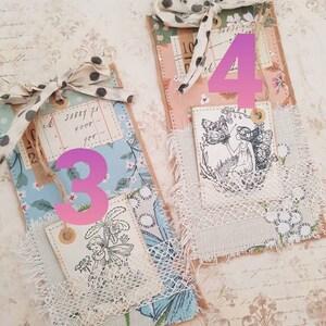 May include: Two handmade paper tags with a vintage aesthetic. Each tag features a different design with a fairy and floral motifs. The tags are decorated with lace, fabric scraps, and twine. The numbers 3 and 4 are written on the tags.
