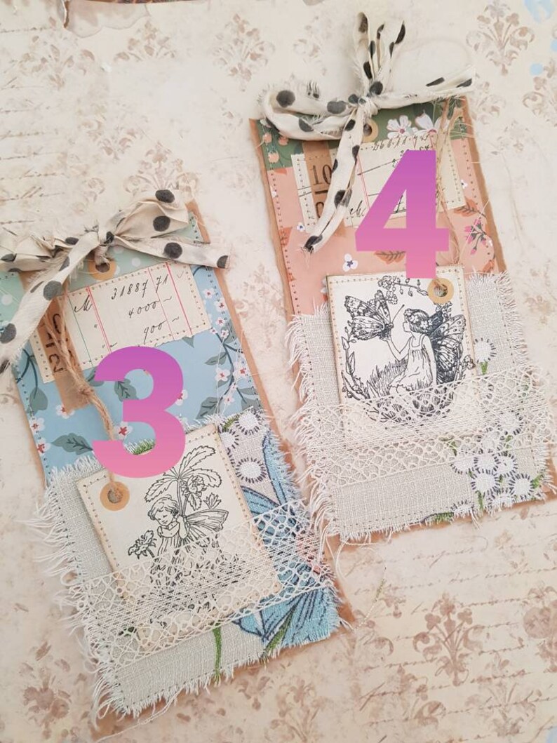 May include: Two handmade paper tags with a vintage aesthetic. Each tag features a different design with a fairy and floral motifs. The tags are decorated with lace, fabric scraps, and twine. The numbers 3 and 4 are written on the tags.