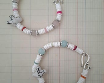 May include: Two white shell bracelets with silver clasps and a small white bird charm. The top bracelet has pink and white beads, while the bottom bracelet has light blue and white beads.