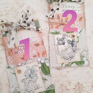 May include: Two handmade fabric tags with a vintage aesthetic. The tags are decorated with lace, floral patterns, and stamped images. The first tag features a stamped image of a plant and the second tag features a stamped image of a fairy.