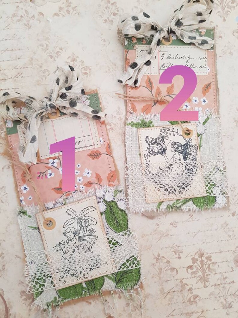 May include: Two handmade fabric tags with a vintage aesthetic. The tags are decorated with lace, floral patterns, and stamped images. The first tag features a stamped image of a plant and the second tag features a stamped image of a fairy.