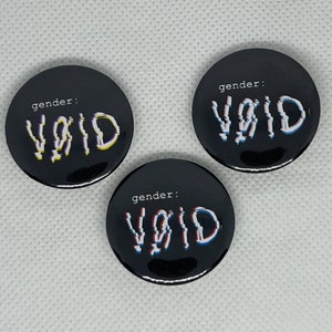May include: Three black round buttons with the text "gender: VOID" in white, with a glitch effect in yellow, blue, and red. The buttons are arranged on a white surface.