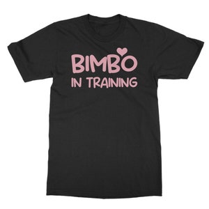 Peut inclure: T-shirt noir avec l'inscription rose "BIMBO IN TRAINING", surmontée d'un cœur rose. Le t-shirt a des manches courtes et un col rond. Le tissu semble doux et confortable.