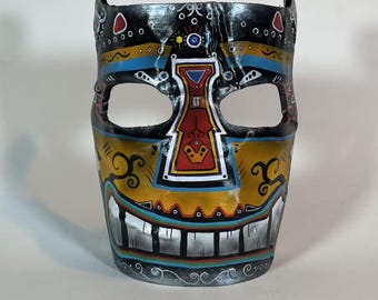 Könnte beinhalten: Eine schwarz, gelb, rot, blau und weiß bemalte Maske mit einem breiten Grinsen. Die Maske hat ein geometrisches Design und erinnert an eine Stammesmaske.