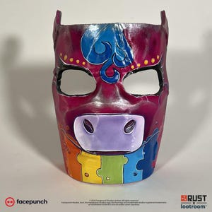 Op de afbeelding: Een roze en paarse koe masker met een regenboogontwerp en een blauwe swirl op het voorhoofd. Het masker heeft ooggaten en een neusgat.