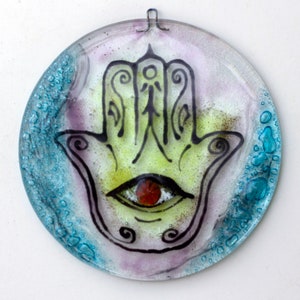 May include: A round glass ornament with a blue, green, and purple background. A black and white hamsa hand with a red eye is in the center of the ornament.