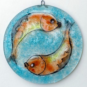 May include: A circular glass plate with a blue background and two orange and yellow fish painted on it. The fish are facing each other and their tails are intertwined. The plate has a textured surface with small bubbles.