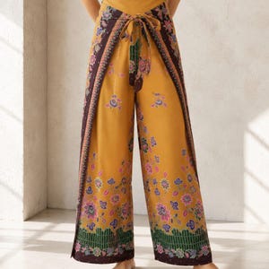 May include: High-waisted wide-leg trousers in a mustard yellow hue featuring a wrap-style front panel with a tie closure. The fabric is adorned with a dark purple and multi-colored floral pattern, including pink and blue accents, with a green grid border.