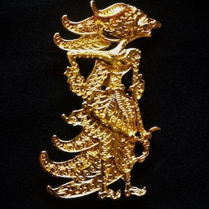 Puede incluir: Un adorno de metal dorado que representa una marioneta tradicional de sombras javanesa, conocida como wayang kulit. La marioneta está intrincadamente detallada con patrones ornamentados y una túnica fluida.