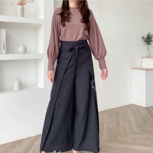 May include: A woman is wearing a long-sleeved, mauve blouse with a black wrap-style pant. The pants have a wide leg and a tie at the waist.