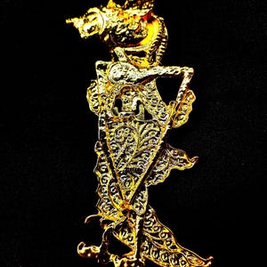以下が含まれることがあります： ワヤン・クリットの影絵人形を模した金色の金属製ブローチ。全身に複雑なフィリグリー（透かし彫り）が施され、尖った頭飾りと、肘を曲げて胴体に添えた細長く伸びたフォルムが特徴的なデザインとなっている。