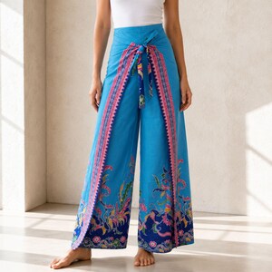 May include: High-waisted wide-leg trousers in bright blue featuring a wrap-style front panel with a tie closure at the waist. The garment displays a colorful floral and leaf pattern in shades of pink, green, and dark blue along the hem and edges.