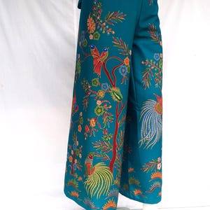 Puede incluir: Pantalón ancho de color azul turquesa con estampado floral y de pájaros. El estampado presenta pavos reales, pájaros más pequeños y flores en tonos rojos, naranjas, amarillos, verdes y azules.