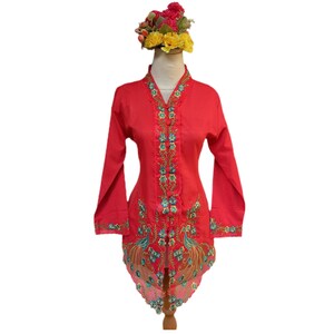 Puede incluir: Una blusa roja vibrante con mangas largas y bordados intrincados en color turquesa y dorado. La blusa presenta un cuello en V y un diseño floral decorativo. Un tocado floral está sobre el maniquí.