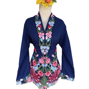 Puede incluir: Una blusa azul marino con mangas largas y bordados florales intrincados. El bordado presenta flores rosas, azules y rojas, junto con dos diseños de pavo real. La blusa se exhibe en un maniquí.