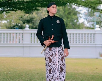 May include: A man wearing a black shirt and a patterned sarong, holding a wooden staff with a red and gold ornament. He is standing on a paved surface in front of a white fence and green grass.
