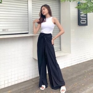 May include: A woman wearing a white tank top and black wide-leg pants with a knotted tie at the waist. She is standing in front of a white building with a window and a sign that says "ONO".