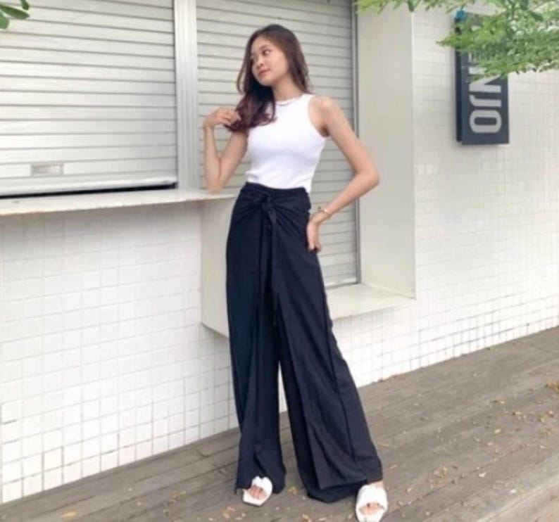 May include: A woman wearing a white tank top and black wide-leg pants with a knotted tie at the waist. She is standing in front of a white building with a window and a sign that says "ONO".