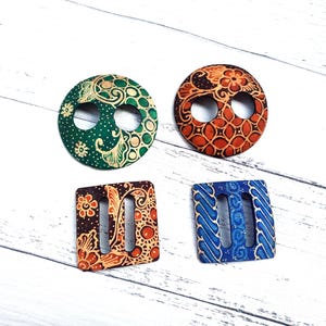 May include: Four wooden belt buckles with intricate batik designs. Two are circular, one green and one orange, and two are square, one brown and one blue. Each buckle features a unique pattern and two openings.
