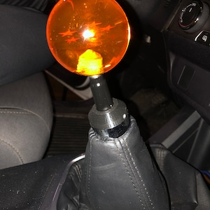 Könnte beinhalten: Orangefarbener und schwarzer Schaltknauf für ein Auto mit einer transparenten Kugel oben. Die Kugel hat ein sternförmiges Design im Inneren.