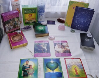 May include: A spread of tarot cards on a white tablecloth with a variety of crystals, candles, and books. The cards include "The Romance Angels Oracle Cards", "Psychic Tarot of the Heart", "Love", "Playfulness", "Trust", "Simply Love", "Love Abounds", and "Strengthening Bonds". The books include "Are Your Own Astrologer" and "The Tarot of the Divine".
