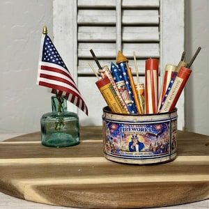 Peut inclure: Une composition patriotique avec un drapeau américain dans une petite bouteille en verre et une boîte de feux d'artifice vintage. La boîte contient divers pétards et fusées, idéale pour les célébrations.