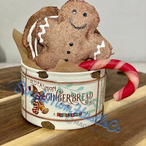 Puede incluir: Un hombre de jengibre decorativo y un bastón de caramelo descansan dentro de una lata blanca. La lata está etiquetada como "Old Fashion Gingerbread Kit" con un tema navideño. El hombre de jengibre es marrón con glaseado blanco y detalles negros.