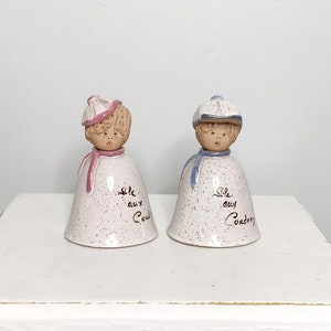 Peut inclure: Deux cloches en céramique avec un glaçage blanc et une finition mouchetée. Les cloches sont en forme de personnes portant des chapeaux et portent les mots "We are so very proud".
