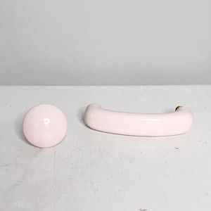 Pode incluir: Dois botões de armário de cerâmica rosa, um redondo e outro em forma de semicírculo.