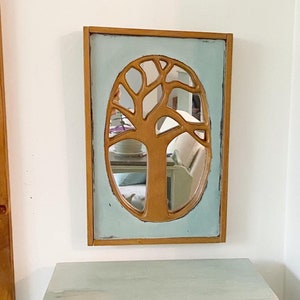 May include: A light blue cabinet with a single drawer and a door. The cabinet has a distressed finish and is decorated with a wooden tree-shaped mirror with a light blue frame.