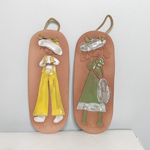Peut inclure: Deux décorations murales en céramique en forme de personnes. La figurine de gauche porte une chemise et un pantalon jaunes et un chapeau blanc. La figurine de droite porte une robe verte et un chapeau blanc.