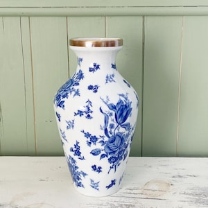 Peut inclure: Un vase en céramique blanc avec un motif floral bleu et un bord doré. Le vase présente un design classique avec des fleurs et des feuilles bleues détaillées. Le vase est posé sur une surface blanche vieillie.