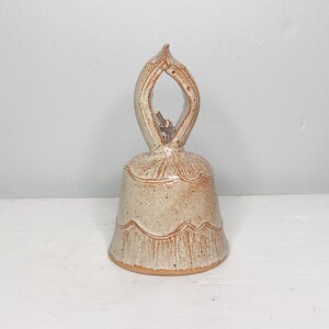May include: A small, white ceramic bell with a brown glaze and a unique, open-work handle. The bell has a scalloped edge and a rustic, handmade look.