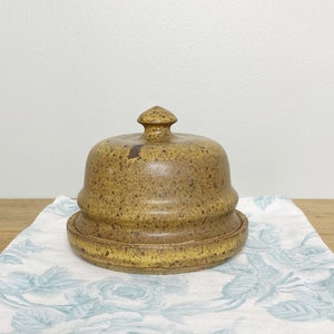 Peut inclure: Un beurrier en céramique brun avec un glaçage moucheté et un couvercle bombé. Le plat est posé sur un tissu à motifs floraux blancs et bleus.