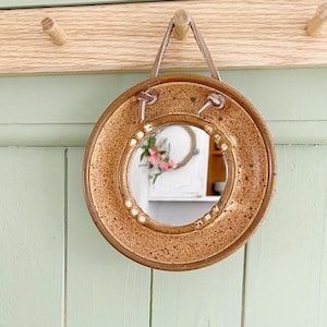 May include: A circular mirror with a speckled brown frame, suspended by a leather strap. The frame is decorated with small, pearl-like studs. It hangs on a light green, panelled wall with a wooden peg rail. The mirror reflects a floral arrangement.